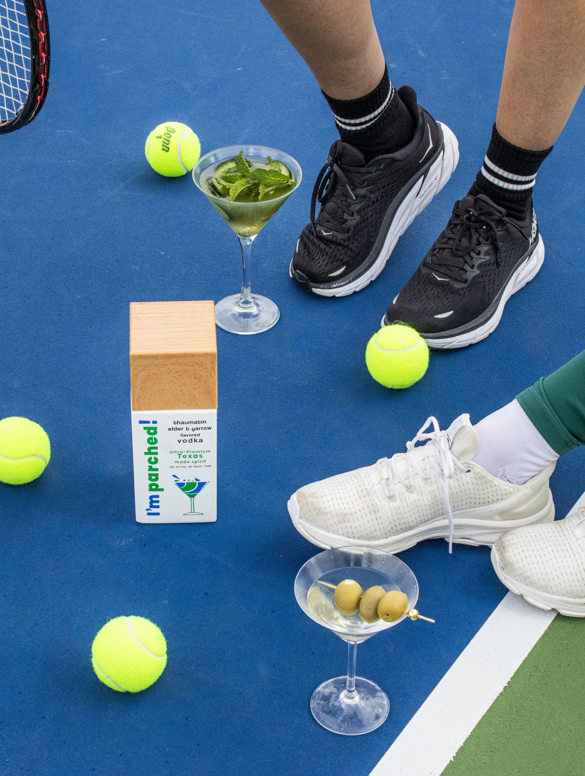 Two people stand on a tennis court with martinis, tennis balls, and a bottle of I’m parched! vodka made for the active lifestyle