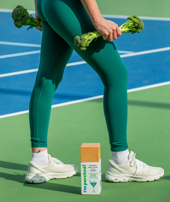 Person holds broccoli like dumbbells on a tennis court with a bottle of ultra premium superfood I’m parched! vodka placed by their fee